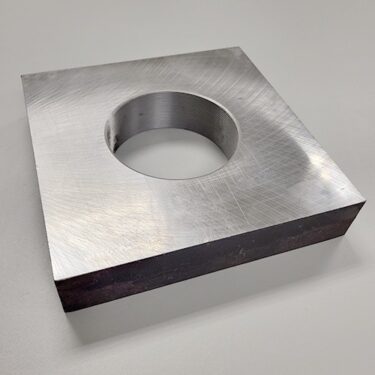 A square metal plate with a threaded central hole, resting on a rough concrete surface.